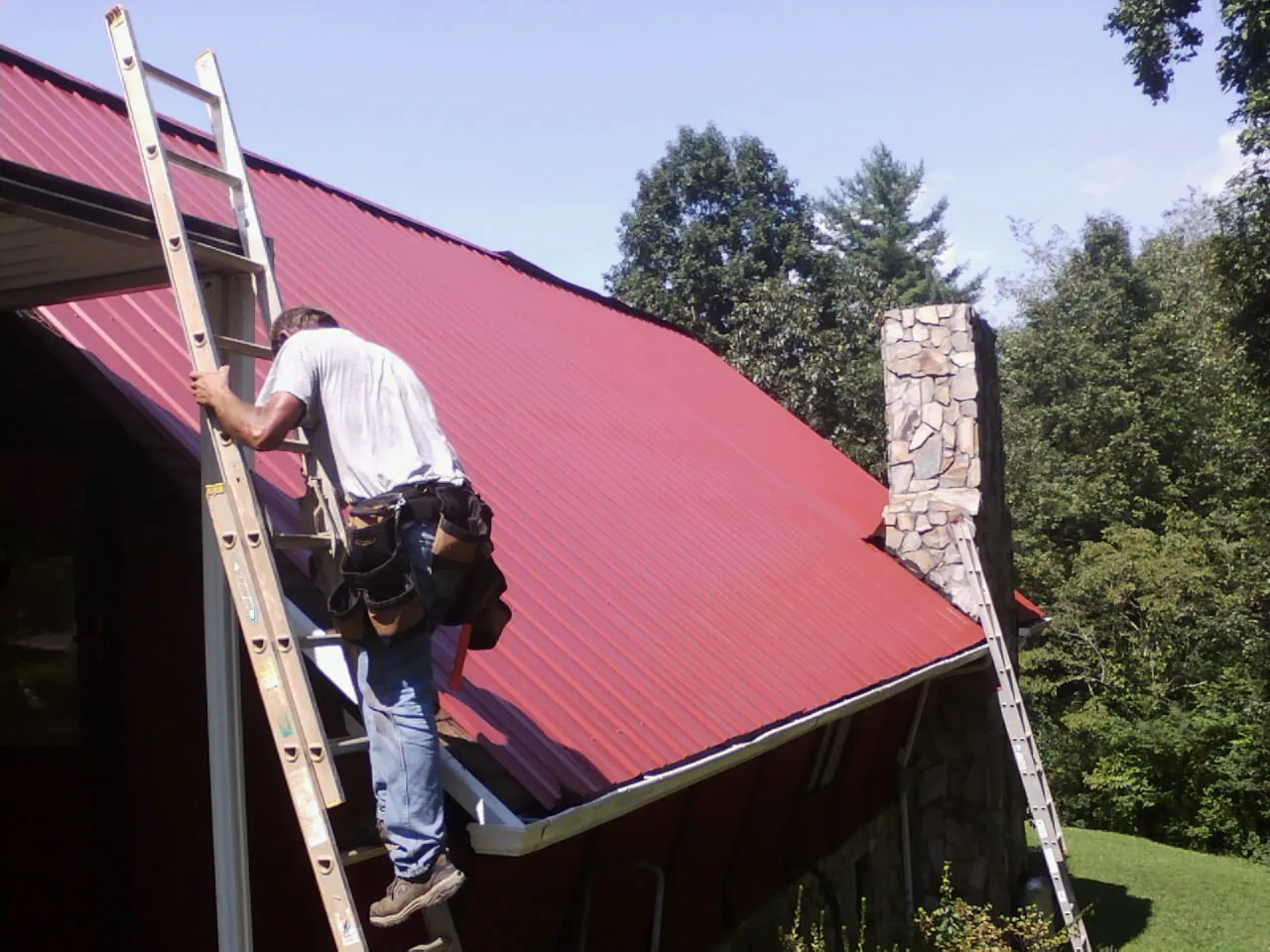 Roofer on a ladder beside a red metal roof and stone chimney — Soffit & Fascia Repair by Roofing Brothers in Ponce De Leon
